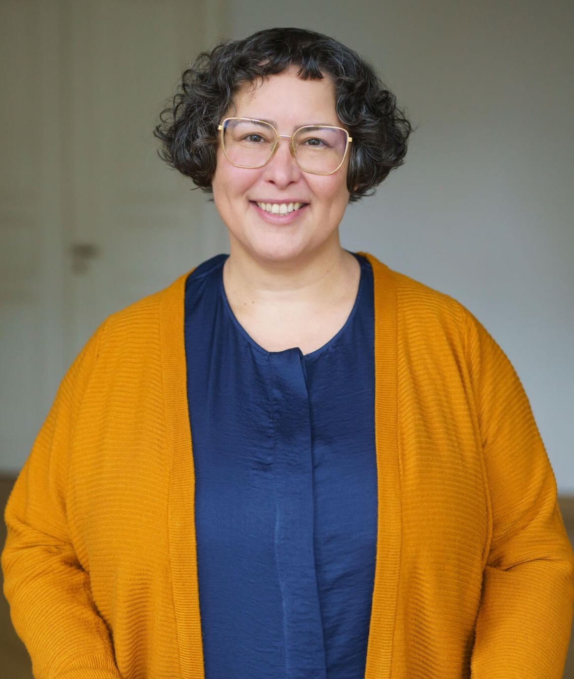 Ira steht lächelnd vor einem hellen Hintergrund. Sie trägt eine blaue Bluse und eine leuchtend gelbe Strickjacke.